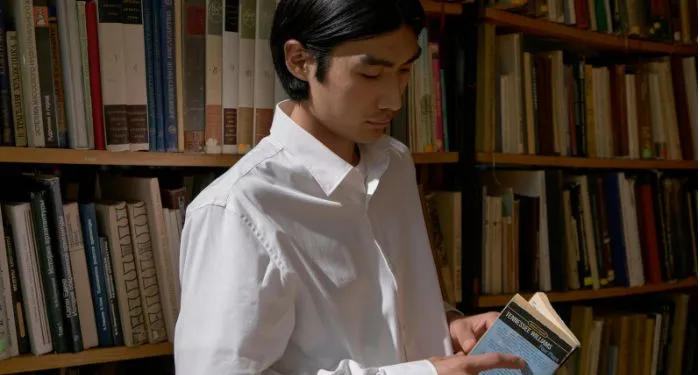 light skinned Asian man reading a book near bookshelves
